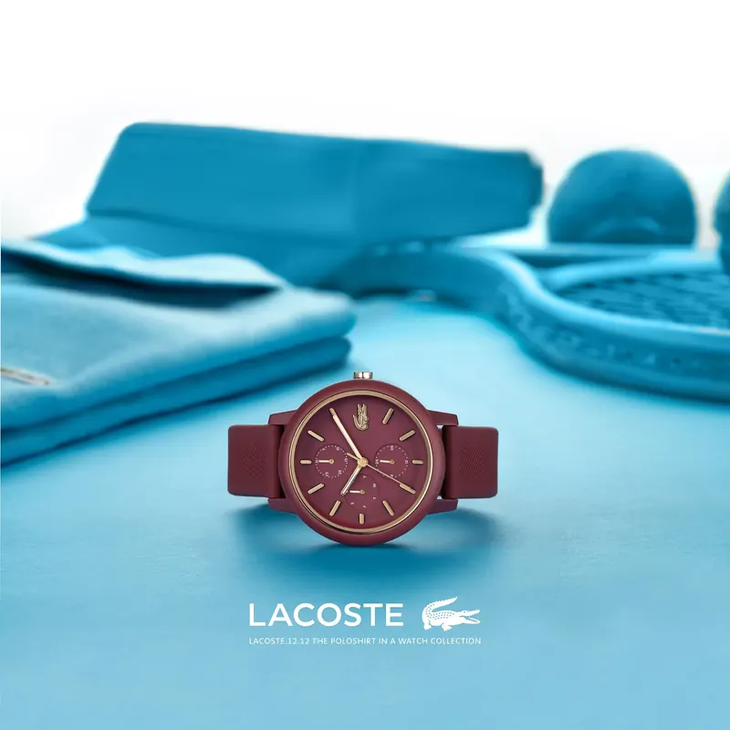 Maroon Lacoste watch on a light blue surface with blue clothing and a tennis racket, Lacoste logo below.