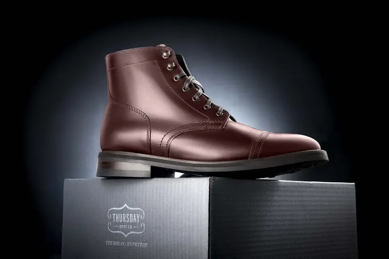 Brown leather cap-toe boot standing on a black Thursday Boot Co. box under a spotlight.
