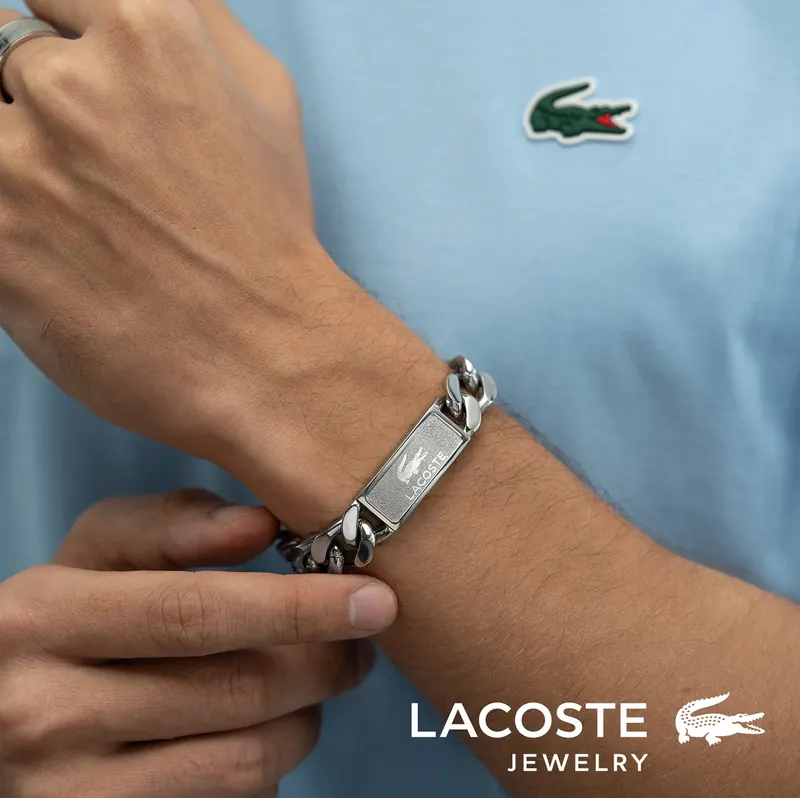 Person in a light blue Lacoste polo fastening a steel ID bracelet, Lacoste Jewelry logo below.