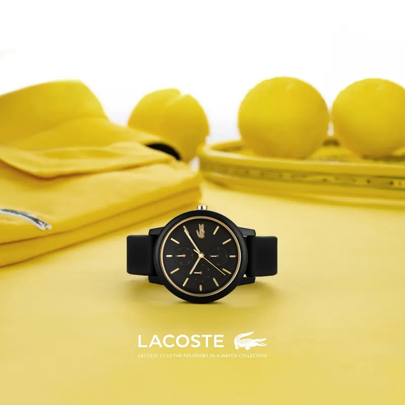 Black Lacoste watch on a yellow surface with yellow clothing and tennis balls, Lacoste logo below.