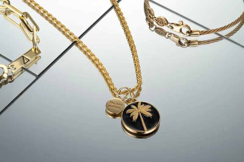 Gold chains and a black enamel palm-tree pendant on angled mirrored surfaces.