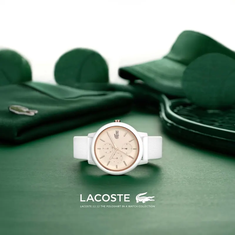 White Lacoste wristwatch with a beige dial on a green surface, surrounded by green clothing and a tennis racket.
