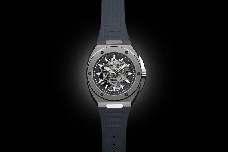 Steel skeleton watch with a dark rubber strap lit by a single soft pool of light on black.