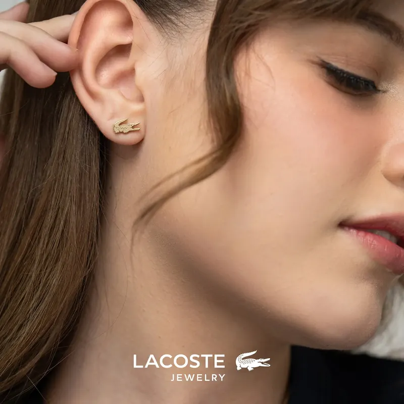 Woman in profile touching her ear, wearing a small gold crocodile stud, Lacoste Jewelry logo below.