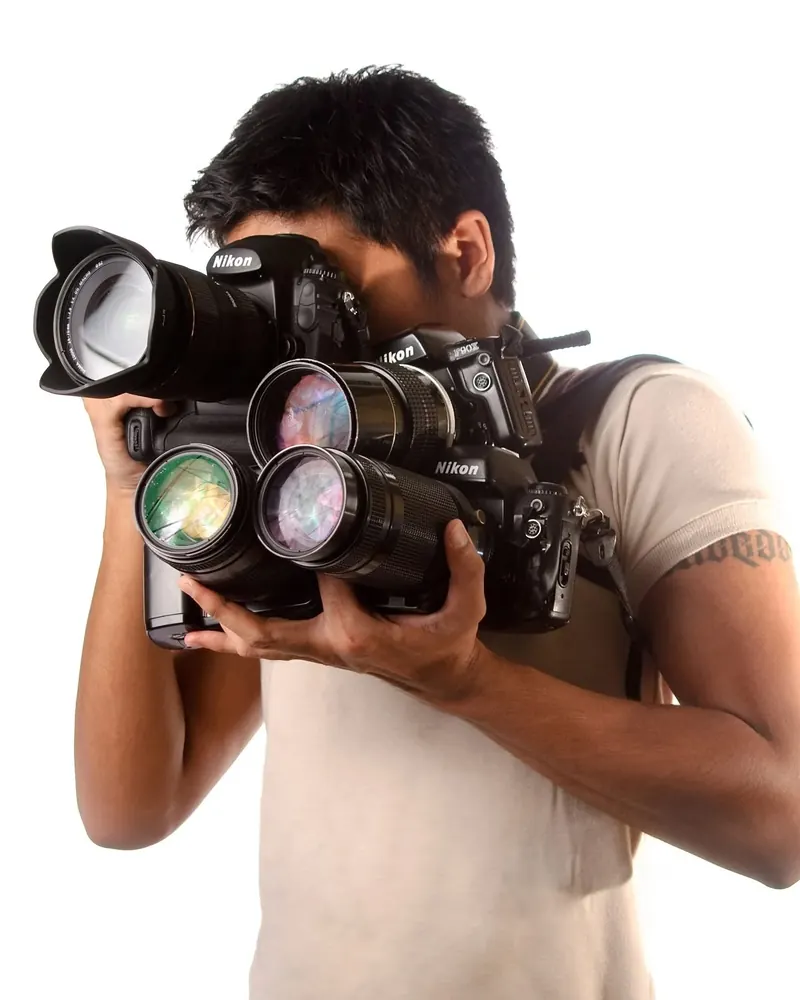 Laya Gerlock holding three Nikon cameras with long lenses, aiming one at the viewer, against a white background.