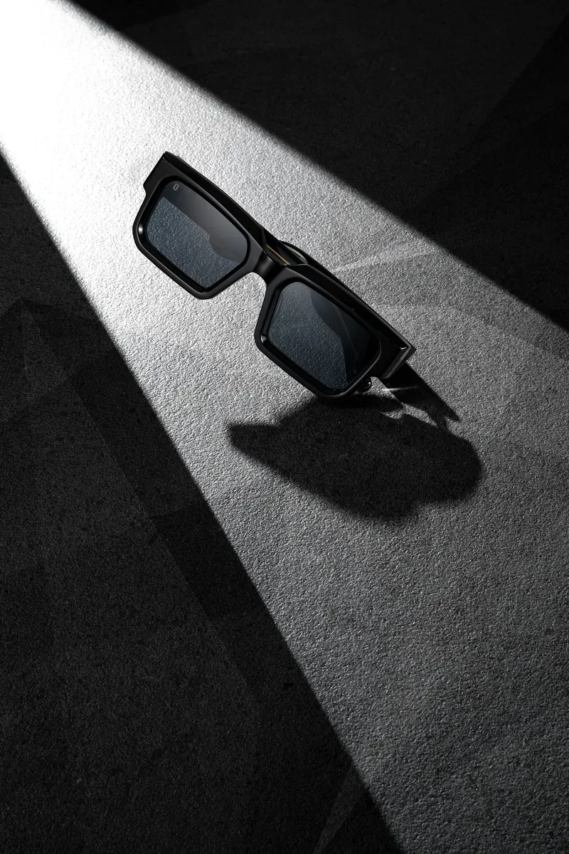 Black square sunglasses casting a sharp shadow in a diagonal shaft of light on dark grey.