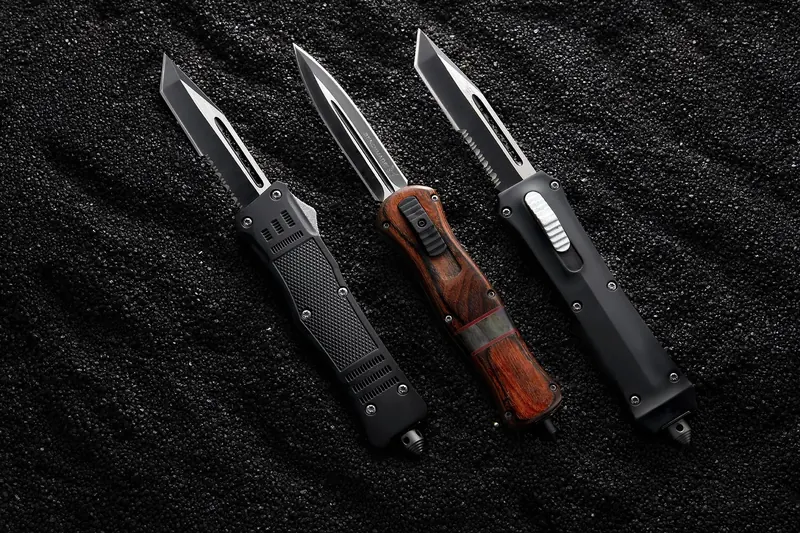 Three automatic knives with black and red handles on black sand.