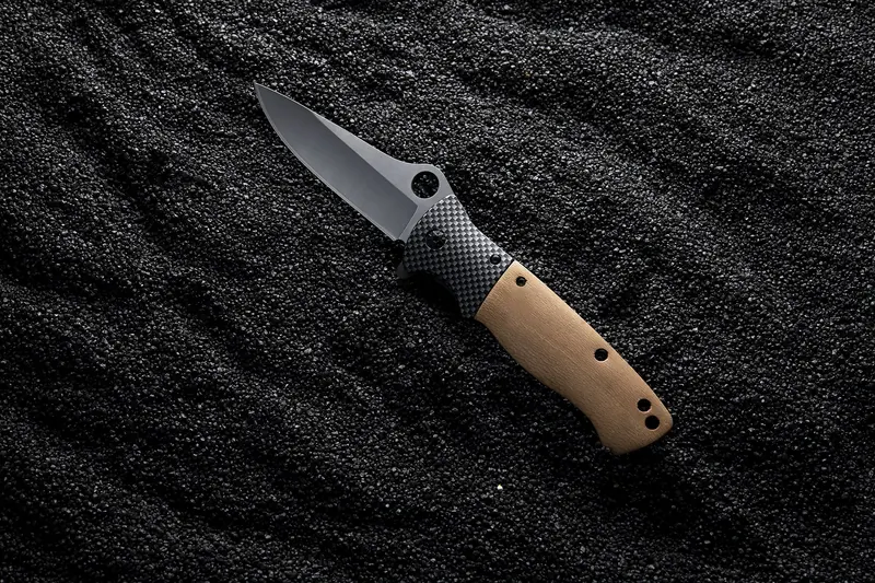 Folding knife with a tan handle and black blade lying on black sand.