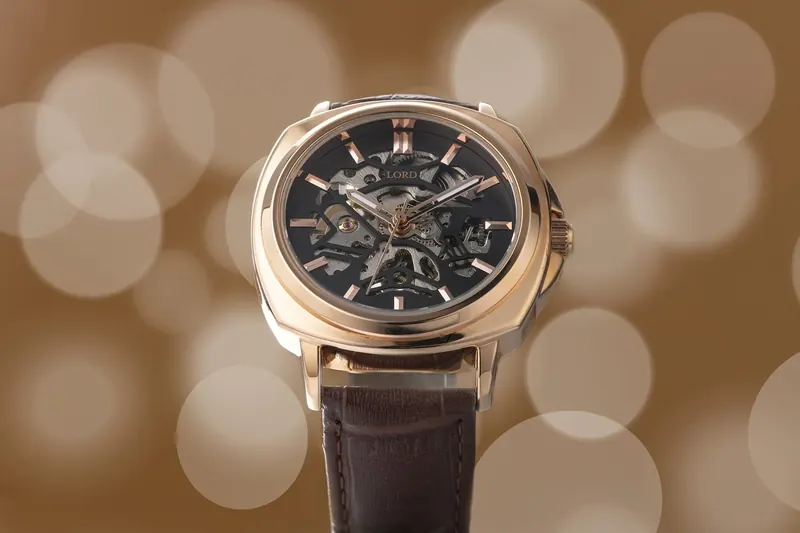 Rose-gold skeleton watch on a brown leather strap in front of warm out-of-focus lights.