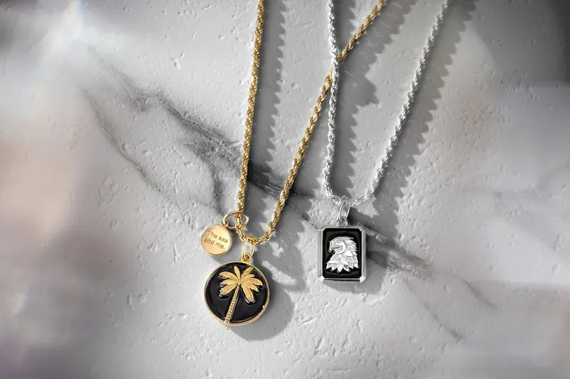 Two pendants, a palm tree and an eagle, on gold and silver chains over white marble.