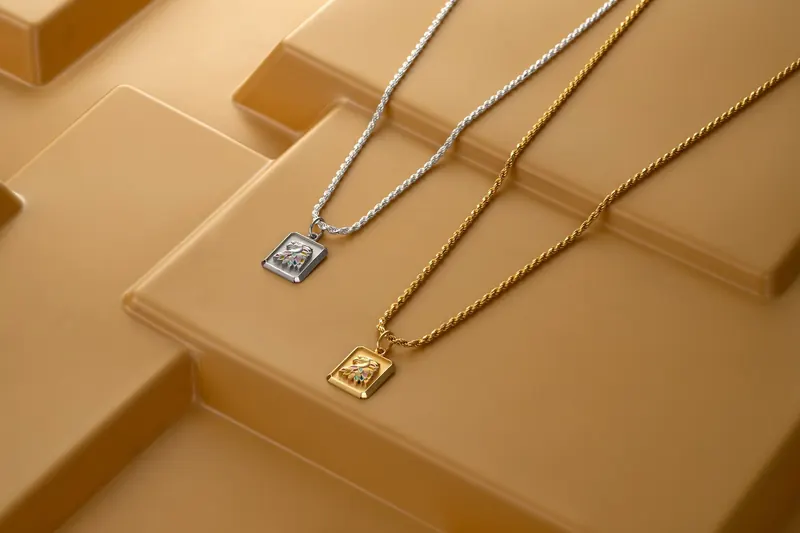 Two tag pendants on gold and silver chains laid over stepped gold-colored blocks.