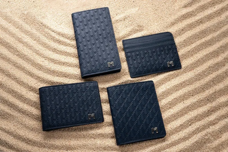 Four navy woven-leather wallets and card holders on raked sand.