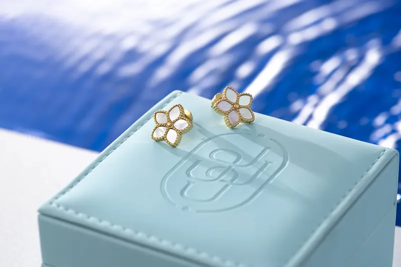 Pair of white flower stud earrings on an embossed pale blue box in front of rippling blue water.