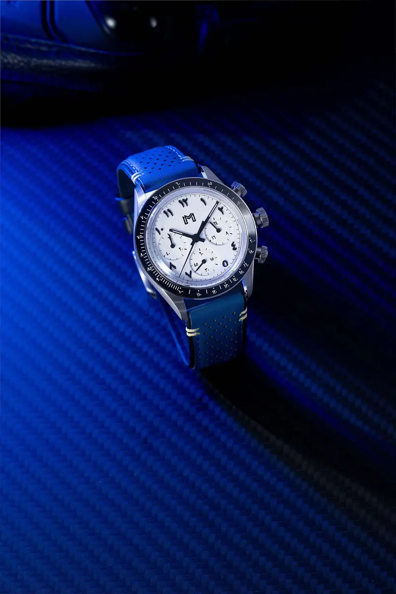 Wristwatch with a white dial, three subdials, black bezel and blue perforated strap on a dark blue textured surface.