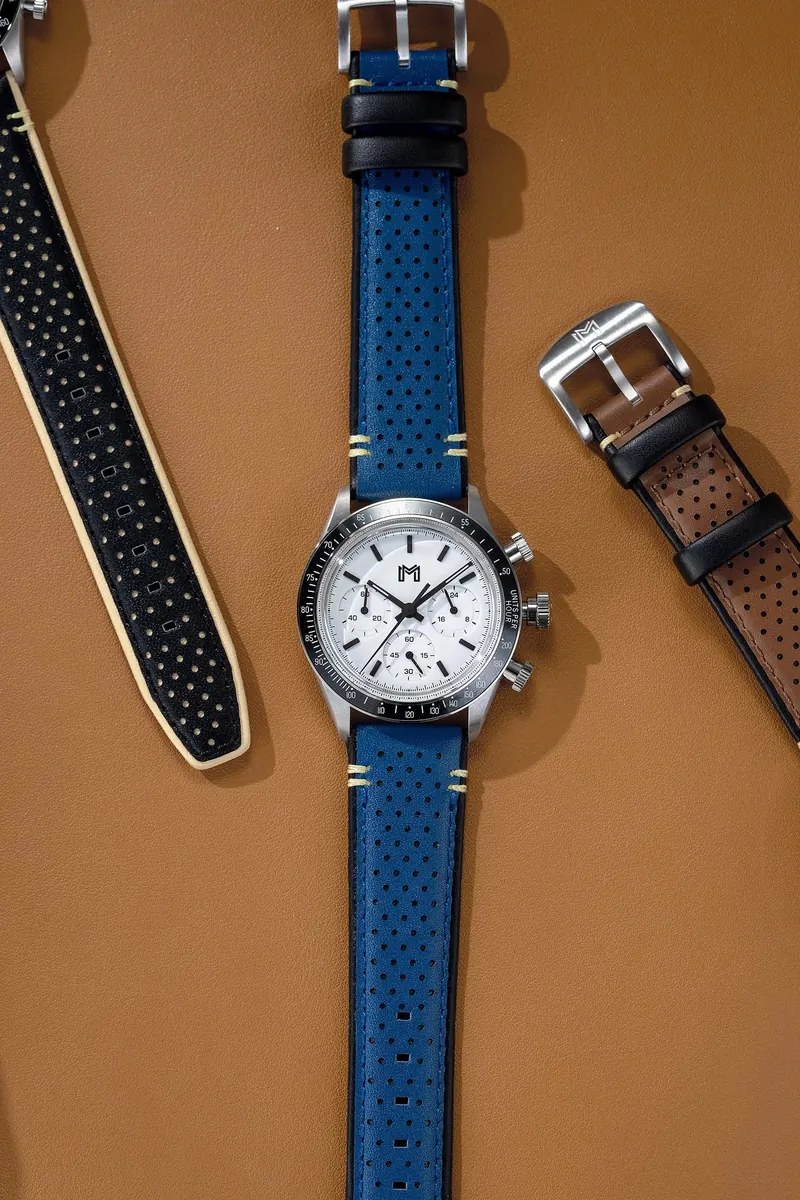 Wristwatch with a white dial and blue perforated leather strap, with two additional straps on a tan background.