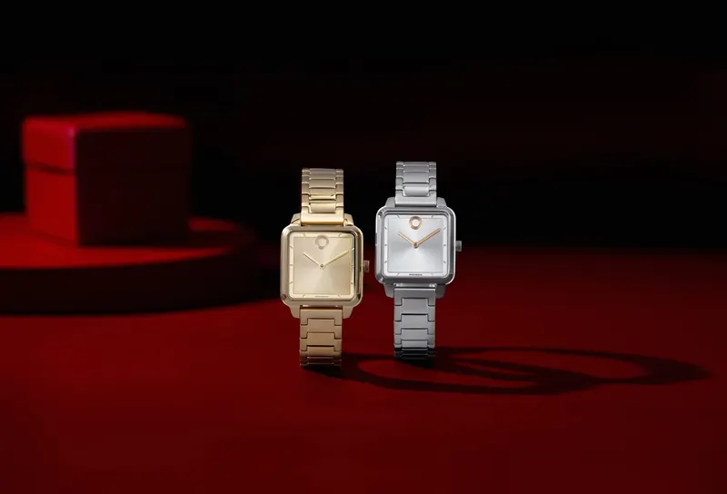 Two square Movado watches, one gold and one steel, standing on a red surface with a red box behind.