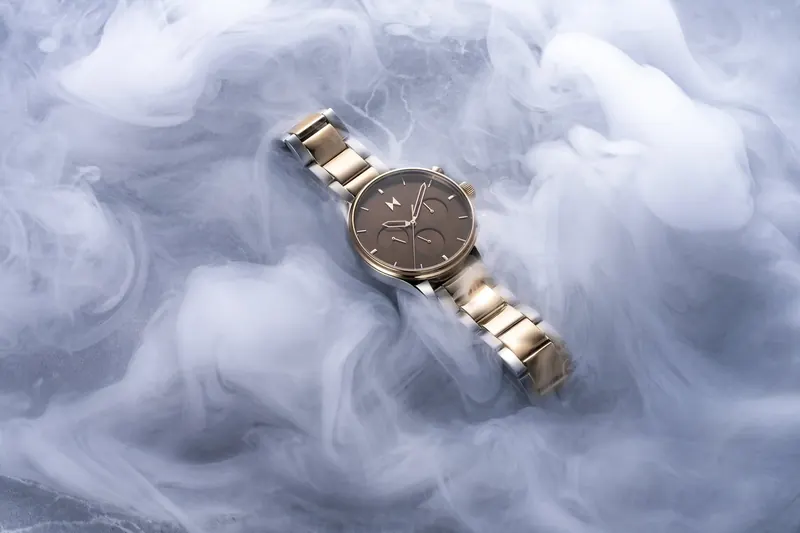 Two-tone MVMT watch lying on soft white smoke against a pale blue background.