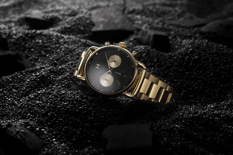 Gold MVMT chronograph with a black dial resting in coarse black sand.