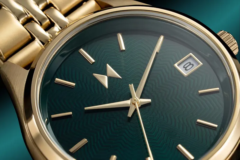 Gold MVMT automatic wristwatch with a dark teal ribbed dial and gold bracelet, shot in macro against a teal background.