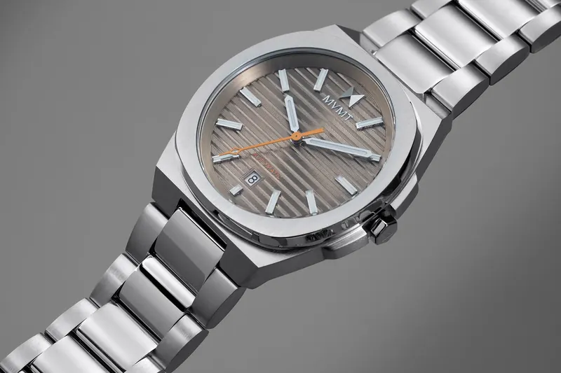 Close-up of a steel MVMT watch with a ribbed taupe dial and integrated bracelet on grey.