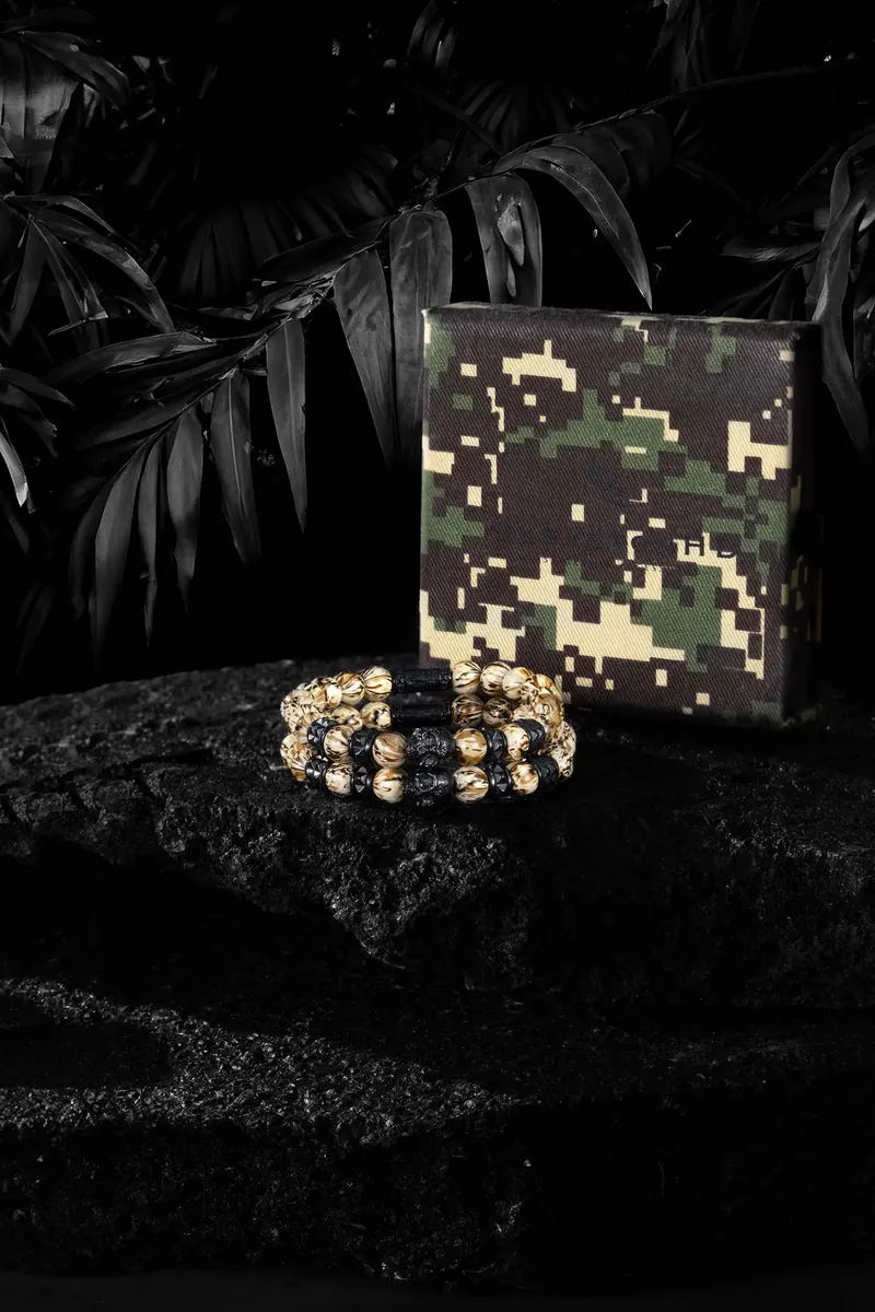 Camouflage box and a black and white beaded bracelet on a textured black surface with tropical leaves in the background.
