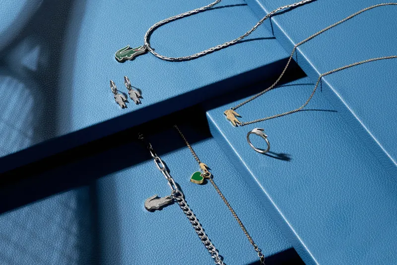 Silver and gold Lacoste necklaces, earrings and rings displayed on stepped blue surfaces under slatted shadow.