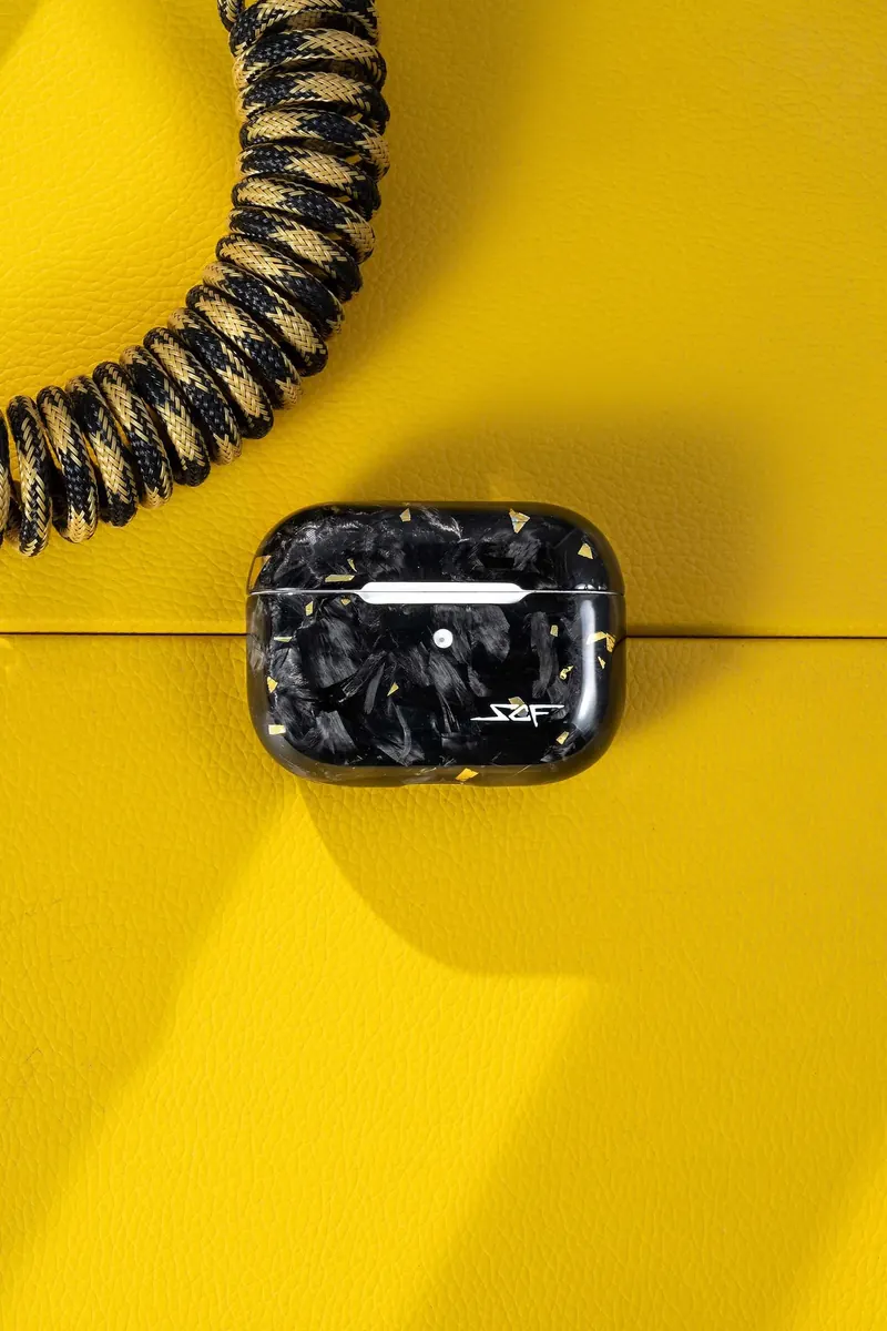 Black and gold patterned wireless earbuds case on a yellow textured surface, with a striped coiled cable.