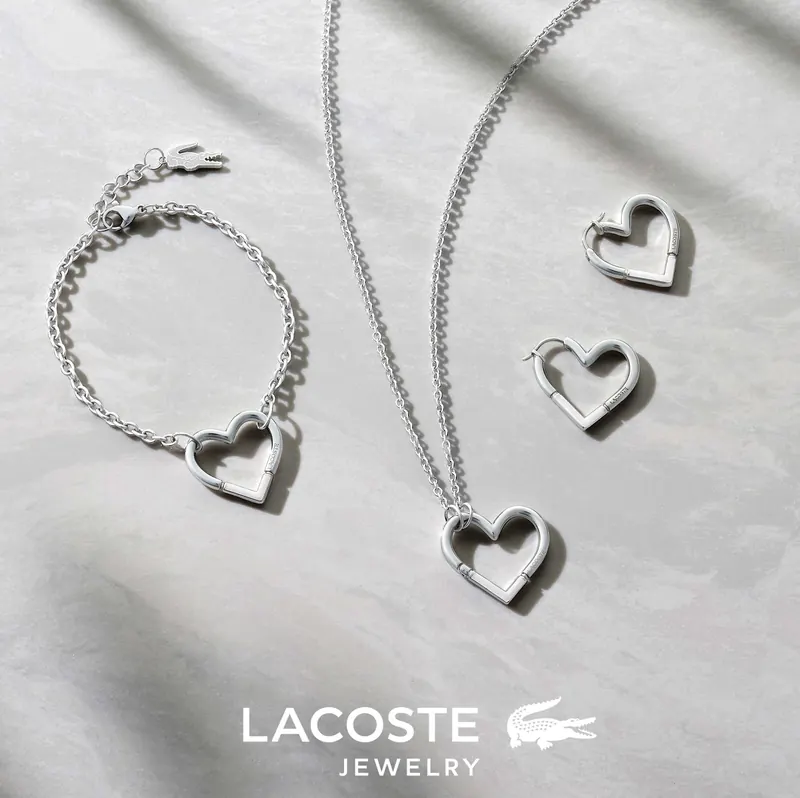 Silver chain necklace, bracelet and heart-shaped earrings on a light surface with the Lacoste Jewelry logo.