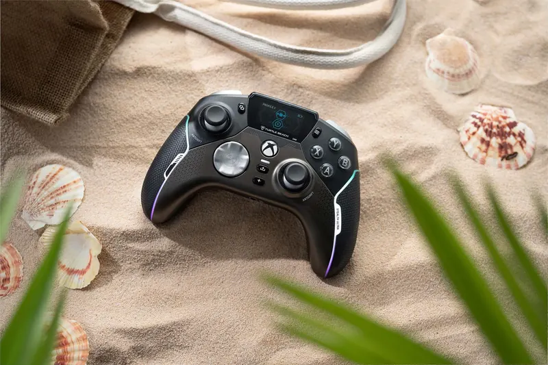 Black Xbox controller on sand with seashells around it, partly covered by green leaves.