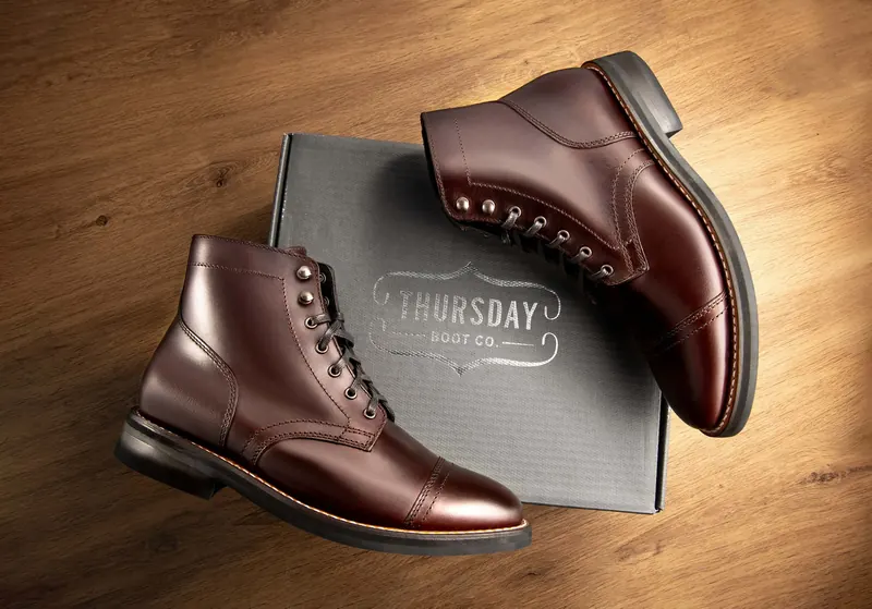 Two brown leather cap-toe Thursday boots resting on a branded grey box on a wooden surface.
