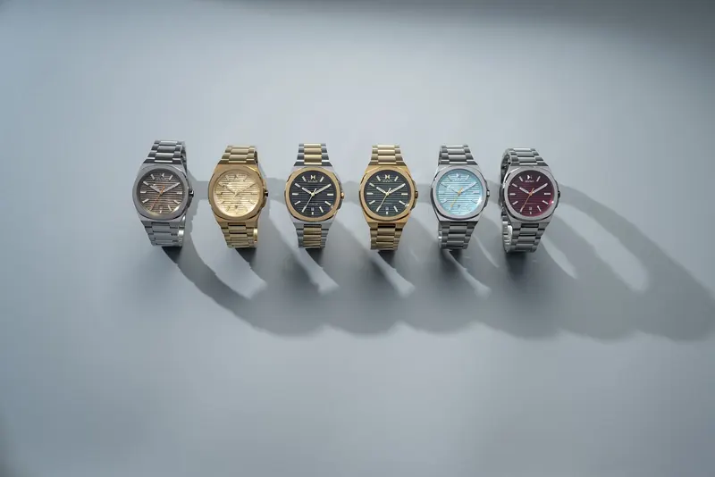 Six steel and gold watches with different colored dials standing in a row on a pale grey surface.