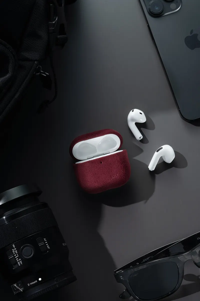 Wireless earbuds and a red case on a dark desk next to a phone, a camera lens and sunglasses.