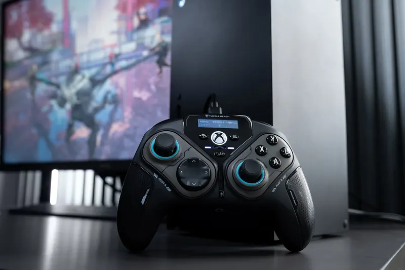 Black game controller on a desk in front of a monitor and a console.