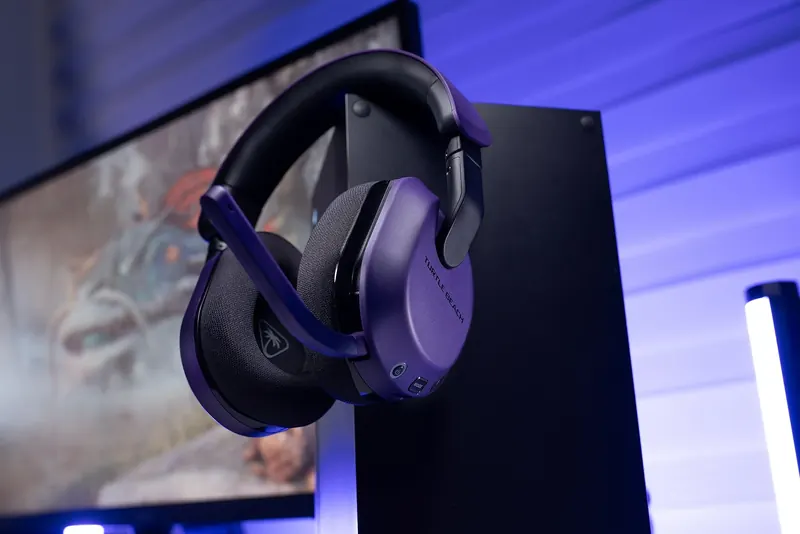 Violet gaming headset hanging on a console beside a glowing monitor.